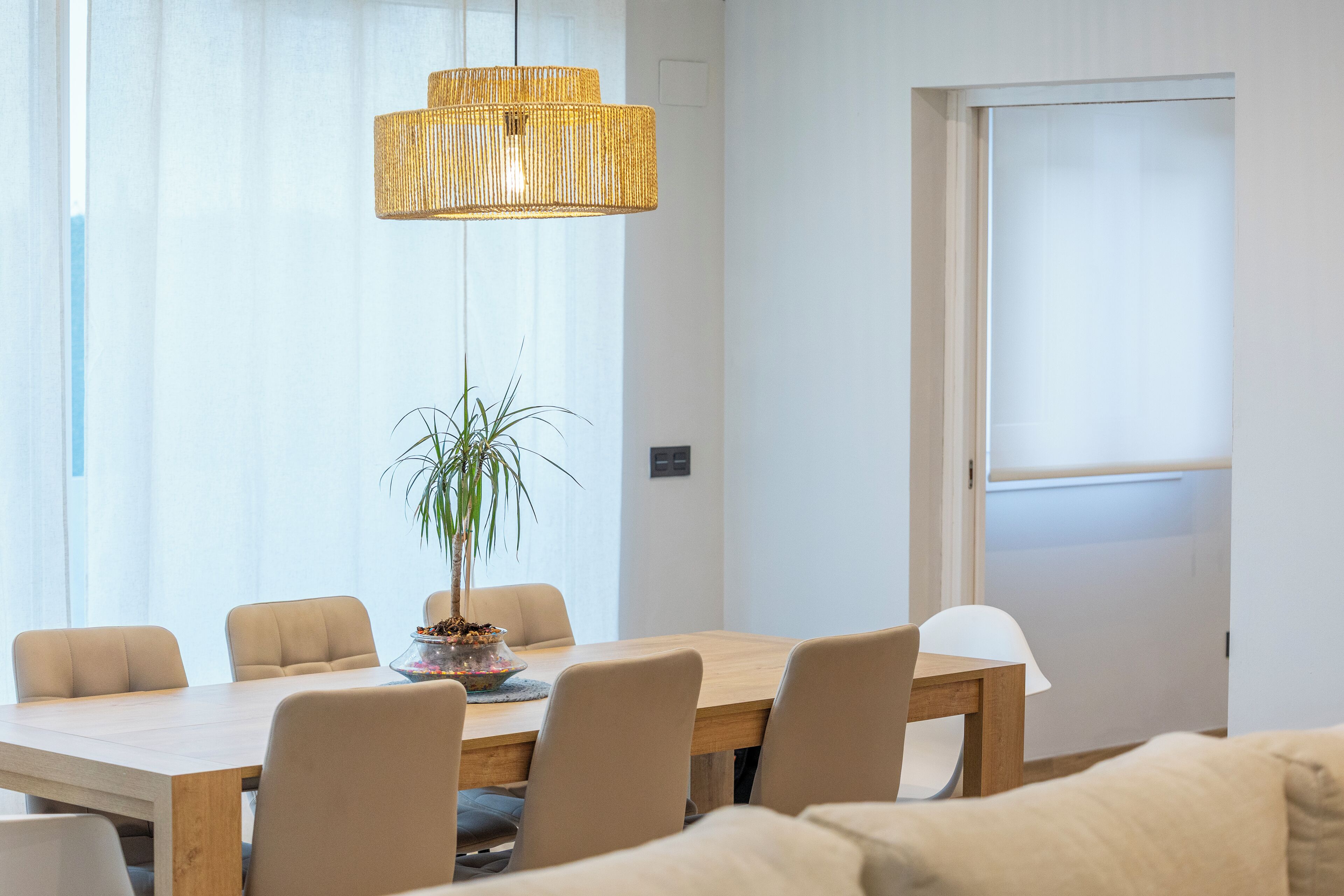 A modern dining area with a wooden table, beige chairs, a potted plant, and a woven pendant light.