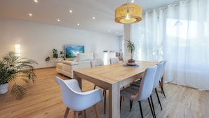 A modern living room with a wooden dining table, white chairs, a sofa, and a large window with sheer curtains. - Bizi Baztan Goienetxea (Elizondo)
