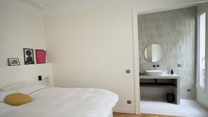 A bedroom with a bed, a wooden herringbone floor, a white wall, a bathroom with a sink and mirror, and a window. - Spacious, Quiet and Elegant Apartment in Paris (Paris)