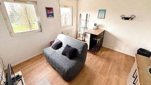 Une pièce avec un canapé gris, une petite table en bois, un radiateur et une fenêtre donnant sur un arbre et des immeubles.