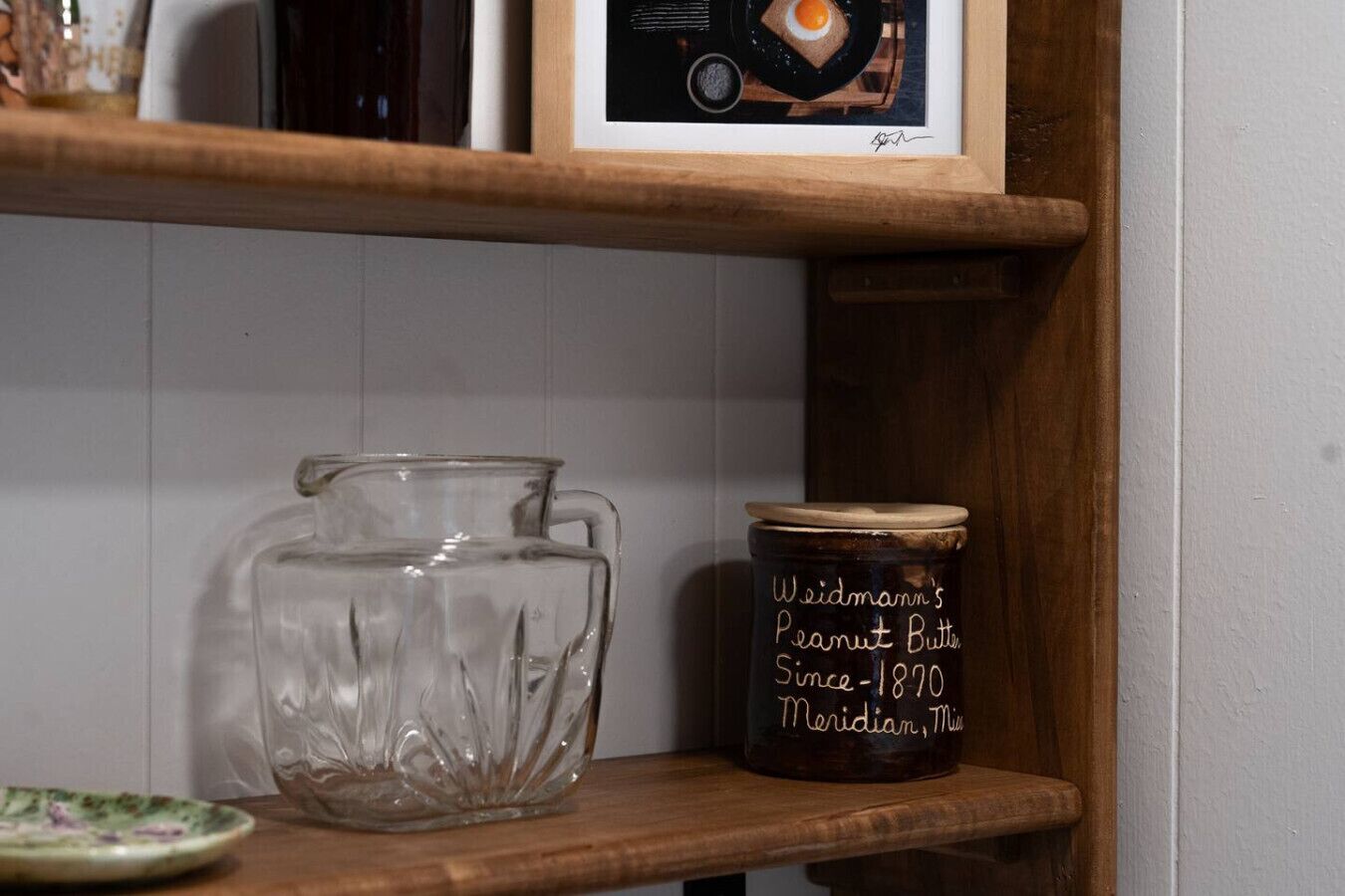 A kitchen shelf with a glass pitcher, a jar labeled 'Weidmann's Peanut Butter,' and a framed picture.
