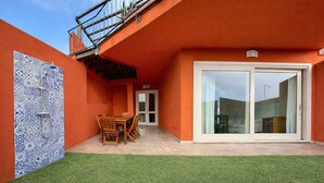 Ein modernes Gebäude mit oranger Fassade, eine Terrasse mit Tisch und Stühlen sowie eine Glastür zu einem überdachten Bereich.