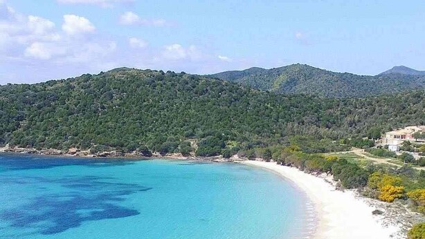 Eine Küstenlandschaft mit klarem türkisblauem Meer, einem Sandstrand und einem bewaldeten Hügel im Hintergrund.