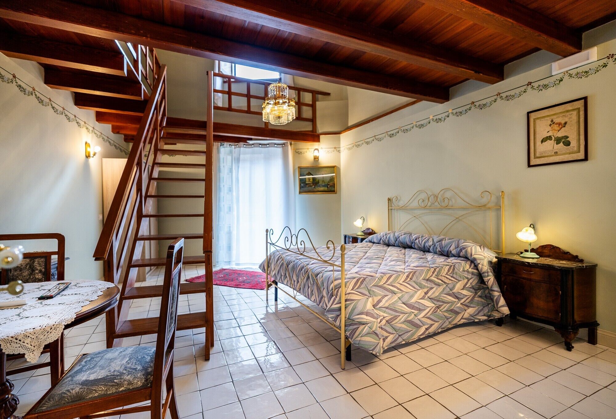 A bedroom with a bed, a wooden staircase, a dining table with chairs, and a chandelier.