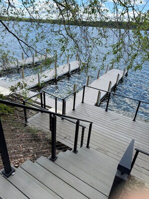 A wooden deck with a metal railing overlooking a body of water, surrounded by trees. - Beautiful 4BR Lake House - New 2026 - Boat Slip - Located on Large Compound (Laconia)