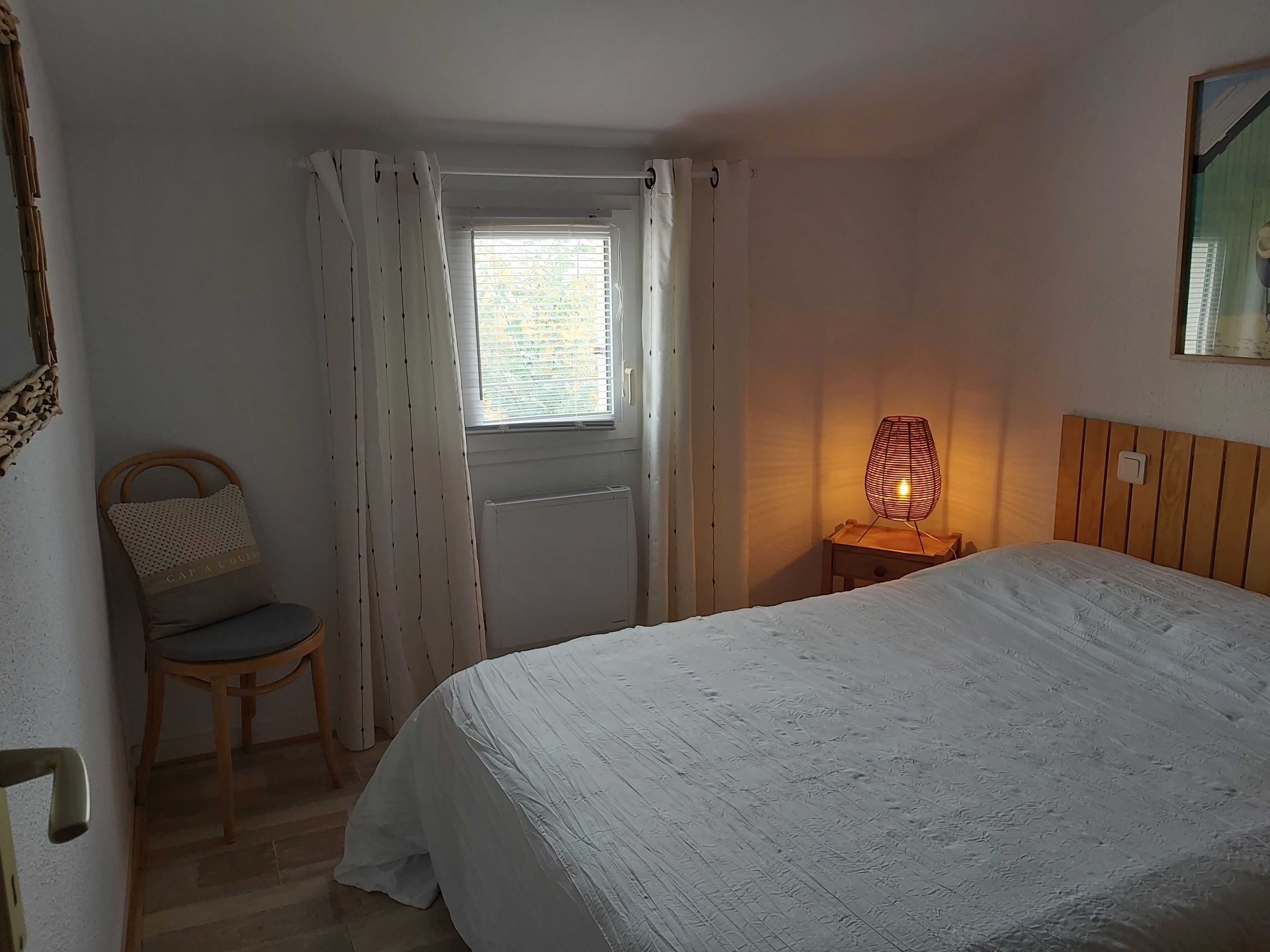 A bedroom with a bed, a chair, a nightstand with a lamp, a window with curtains, and a radiator.