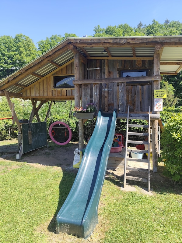 A rustic wooden playhouse with a slide, surrounded by greenery.