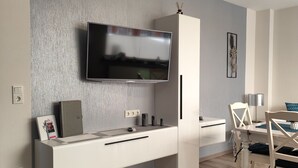 A modern living room with a flat-screen TV mounted on a textured wall, a white entertainment unit with a drawer, a dining area with a wooden table and chairs, and a light-colored floor.
