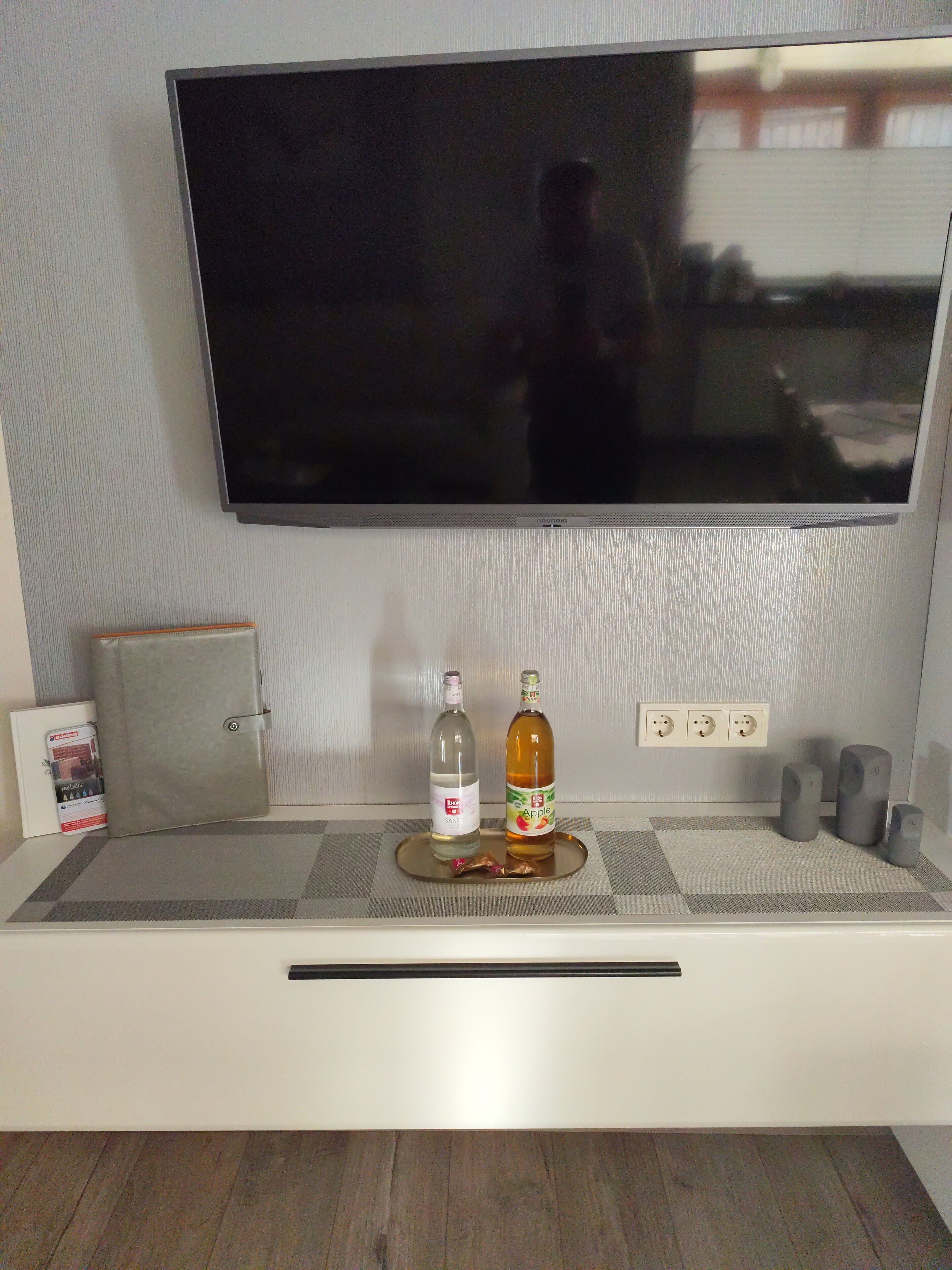 A flat-screen TV mounted on a wall, a small table with a tray holding two bottles, and a power outlet with three plugs.