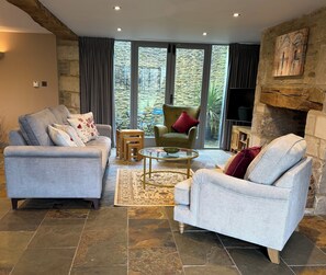 A living room with two grey sofas, a green armchair, a wooden coffee table, and a stone fireplace. - Luxury Cotswold Cottage Can Sleep 8. Secure Garden & Parking.  Walks From Door (Poulton)