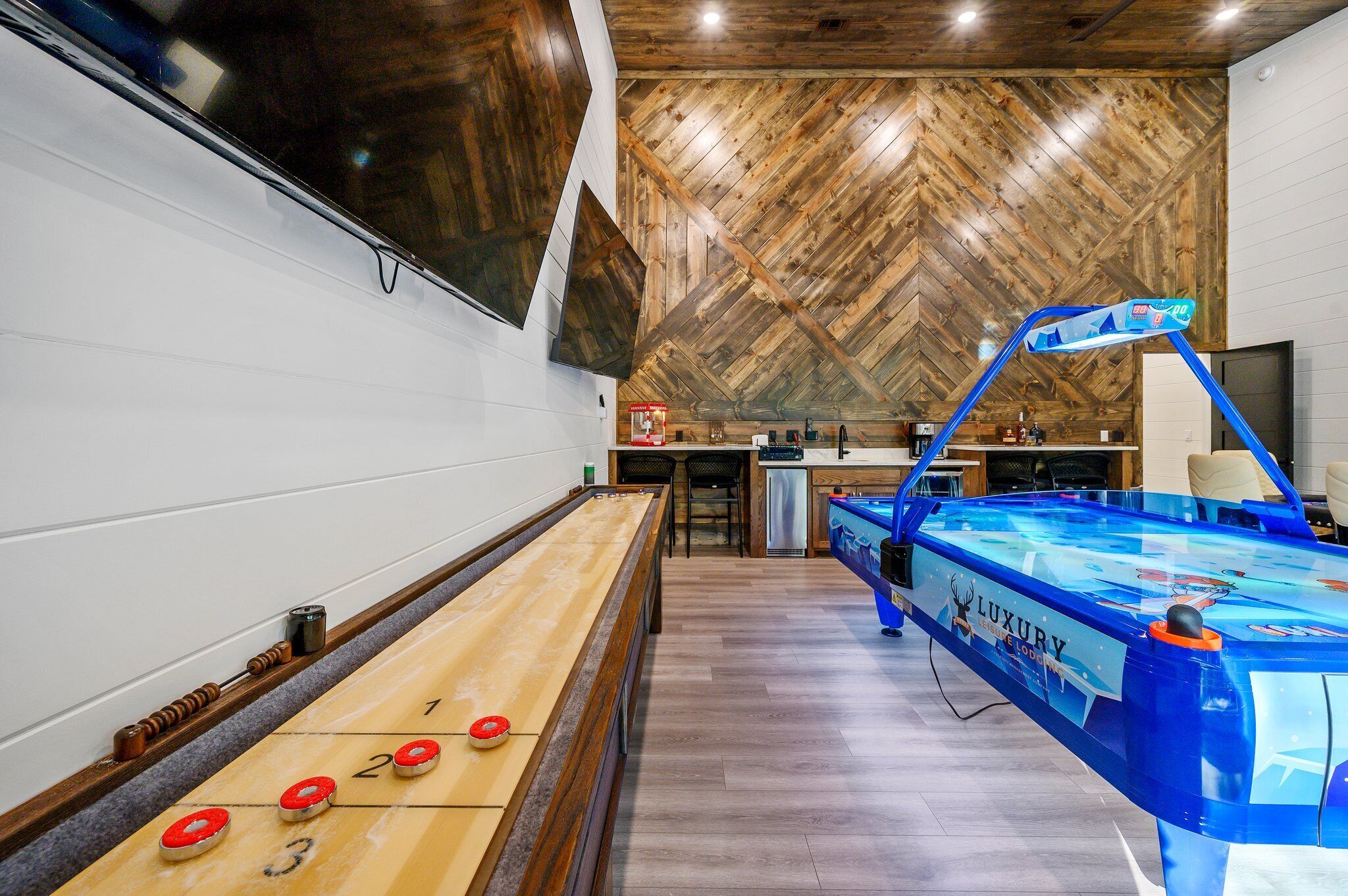 A shuffleboard table, a pool table, and a bar area in a room with wooden ceiling and white walls.