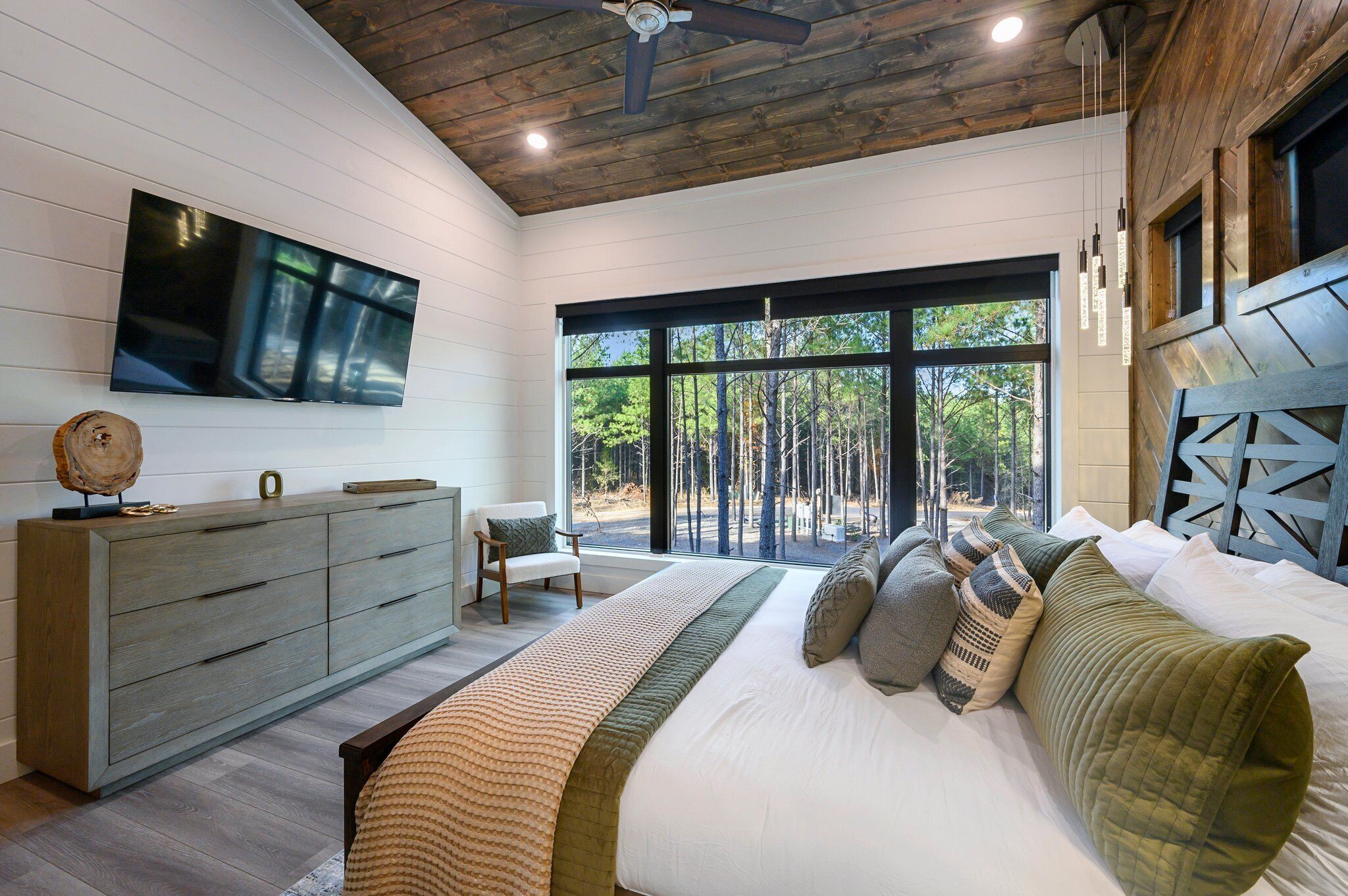 Een slaapkamer met een groot bed, houten plafond, een aan de muur bevestigde televisie en uitzicht op bomen door grote ramen.