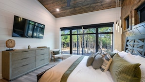 Quarto com uma cama grande, teto de madeira, televisão na parede e vista para árvores por grandes janelas.