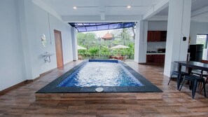 A swimming pool with a wooden deck, a kitchen area, and a view of greenery.