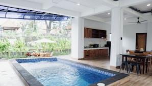 A modern indoor swimming pool with a view of a tropical garden, wooden furniture, and a kitchen area.