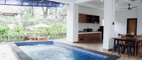 A modern indoor swimming pool with a view of a tropical garden, wooden furniture, and a kitchen area.