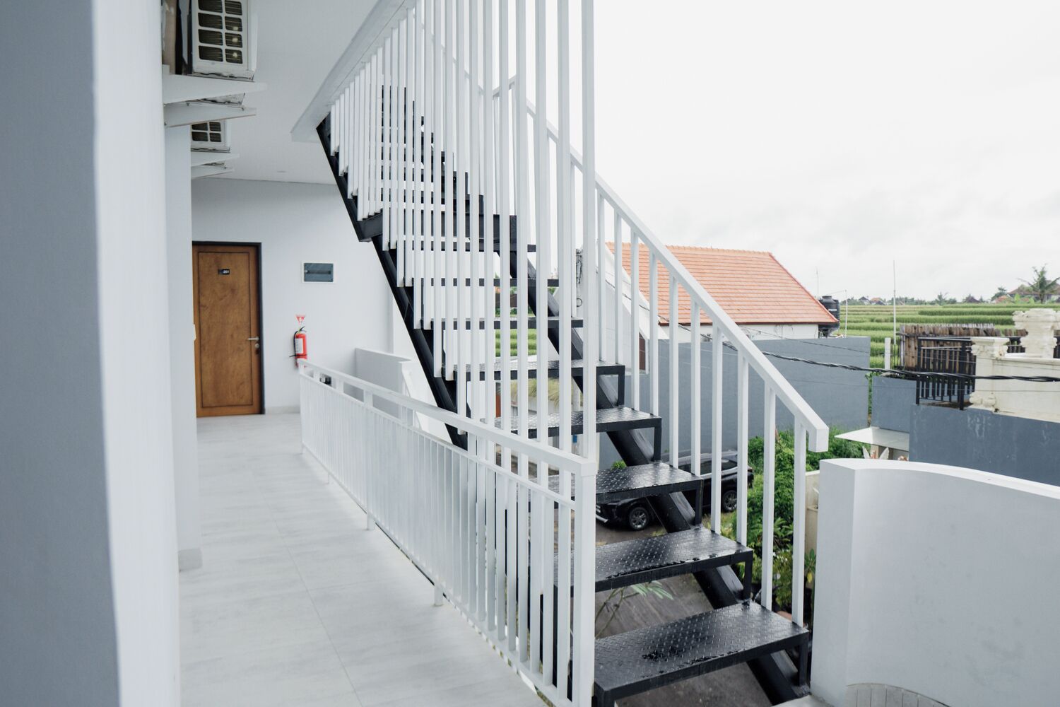 A modern staircase with white railings and black steps, leading to an outdoor area with a view of a red-roofed building and green fields.