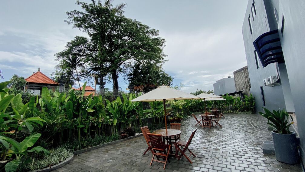 An outdoor patio with tables and chairs, surrounded by lush greenery and a large tree.