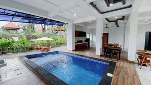 A modern indoor swimming pool with a view of a garden, a dining area, and a kitchen.