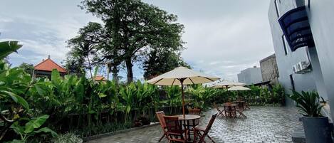 Un espace extérieur aménagé en terrasse, avec des tables et des chaises, entouré d’une végétation luxuriante et, au fond, un bâtiment doté d’un toit rouge.