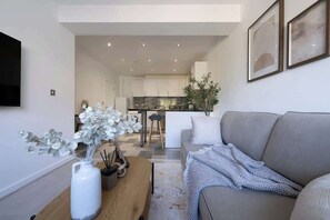 A modern living room with a grey sofa, a wooden coffee table, and a white vase with flowers. A kitchen is visible in the background.