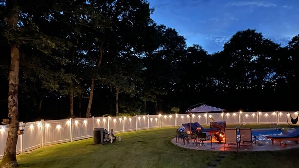 Un patio trasero con piscina, sillas de descanso, una fogata y luces colgantes a lo largo de la cerca.