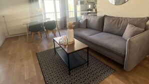 A living room with a grey sofa, a coffee table with a tray, a patterned rug, a round mirror on the wall, and a kitchen in the background.