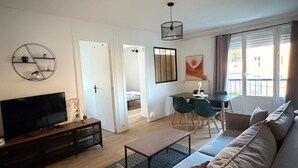 A living room with a sofa, TV, dining table, and chairs. There is a door leading to another room.