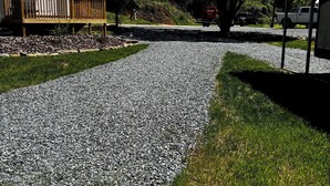 A gravel driveway leading to a house with a wooden deck and a large tree.