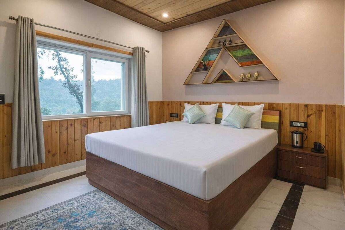 A bedroom with a large bed, wooden paneling, a window with curtains, a nightstand with a kettle, and a wall-mounted shelf with decorative items.
