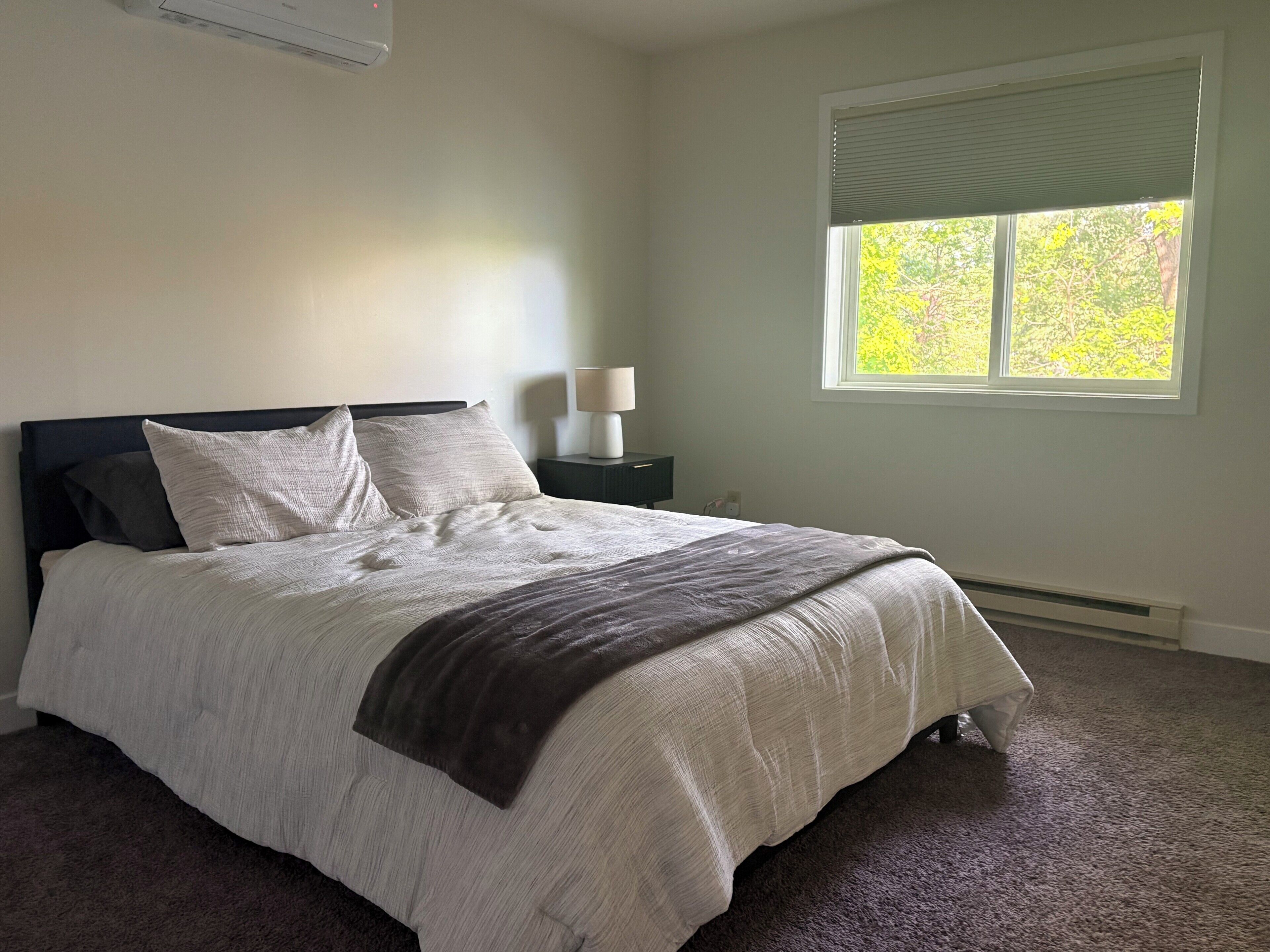 A bedroom with a bed, a nightstand, a lamp, and a window with a view of trees.
