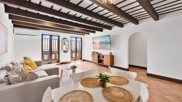 Uma sala de estar moderna com teto alto, vigas de madeira embutidas, um conjunto de sofá branco, uma mesa de jantar com jogos americanos trançados e vista para o exterior através de janelas amplas.