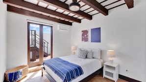 Quarto com cama, mesas de cabeceira, janela e porta que dá para uma varanda.