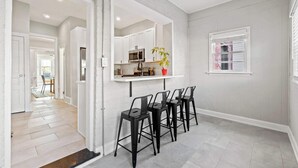 Una cocina moderna con armarios blancos, microondas y una planta. Hay cuatro taburetes negros y una ventana con persianas.