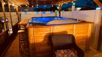 A wooden deck with a hot tub, a wicker chair, and a wooden table.
