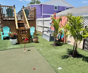 A backyard with artificial grass, a wooden play structure, a purple house, and a checkered fence. - Sunset By The Sea • Entire Property • 4BR/2BA • Pool & Swim (Fort Myers Beach)