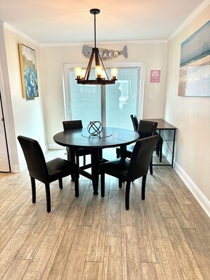 A dining area with a round table, four chairs, a chandelier, and a window with blinds. - Sunset By The Sea • Entire Property • 4BR/2BA • Pool & Swim (Fort Myers Beach)