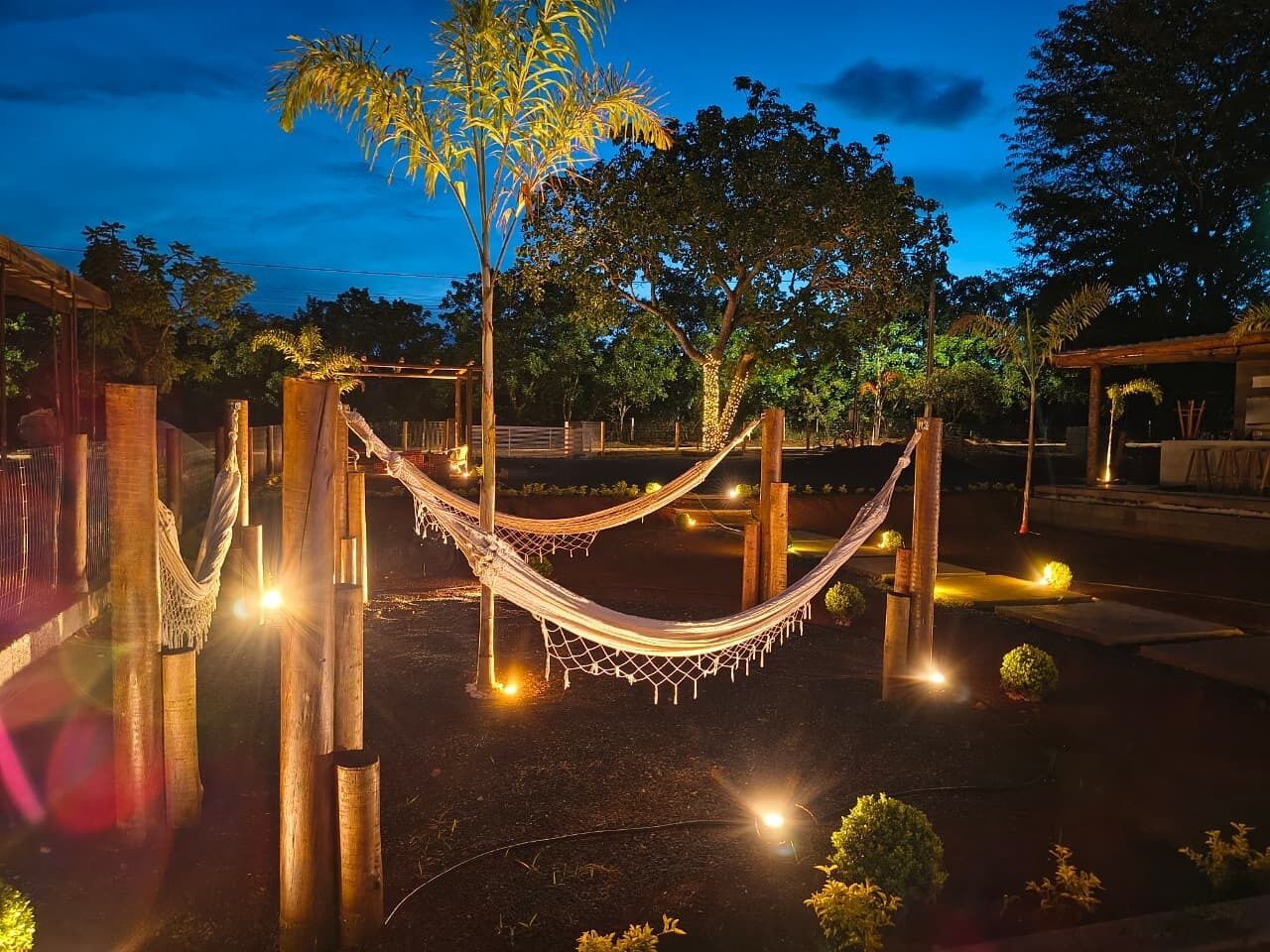 Una hamaca entre postes de madera en un jardín con árboles e iluminación.