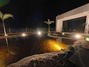 Un área de piscina exterior iluminada de noche, con un edificio moderno y palmeras.