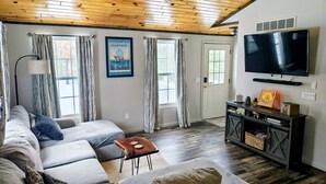 A living room with a wooden ceiling, a large TV, a sofa, a coffee table, and a wall-mounted shelf with decorative items. - Beautiful private home located in Lewiston Michigan. (Lewiston)