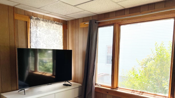 Ein Schlafzimmer mit Fernseher auf einer Kommode, einem Fenster mit Vorhängen und einem Bett mit gesteppter Bettdecke.
