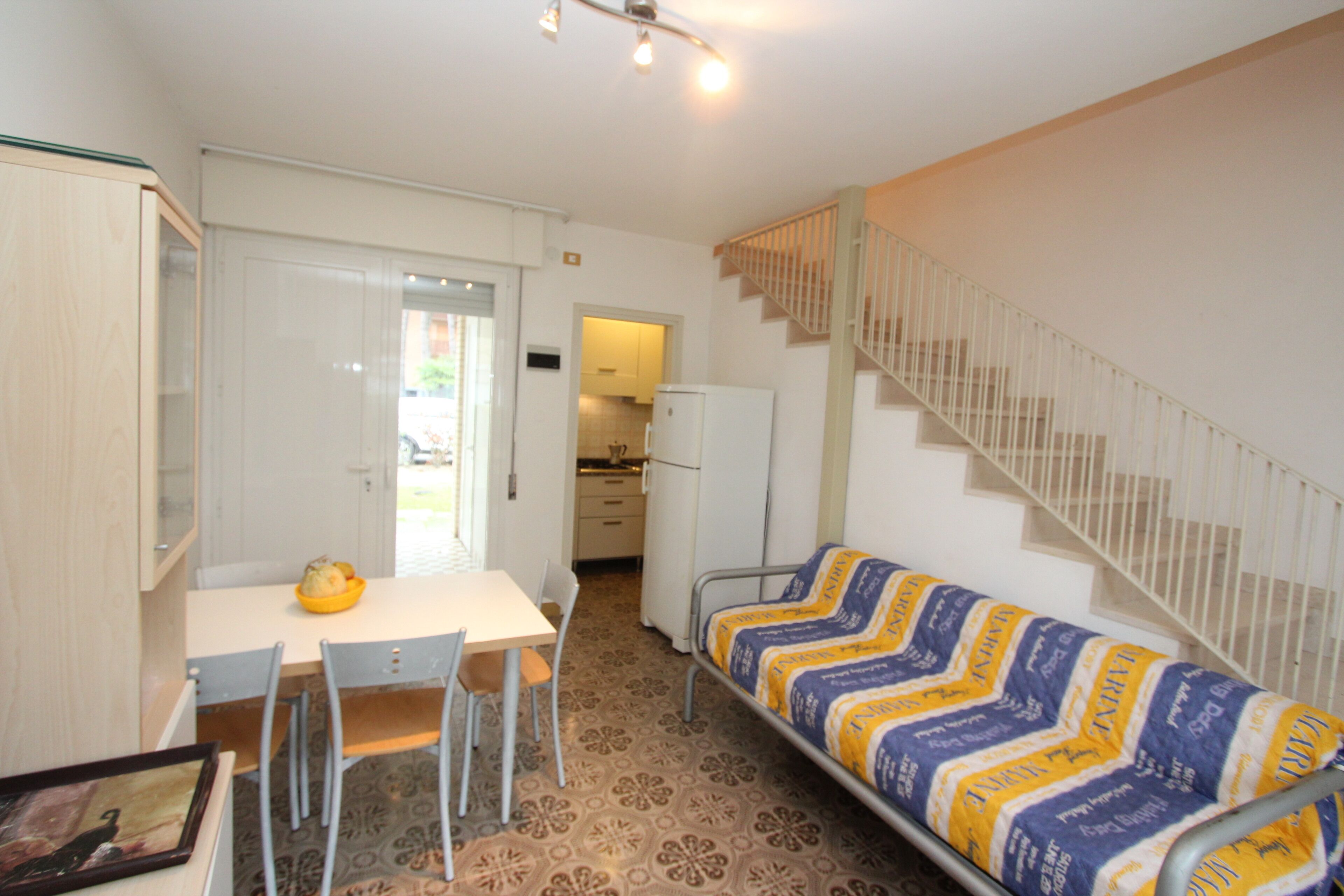 A compact living space with a dining area, a sofa, a staircase, and a kitchen in the background.