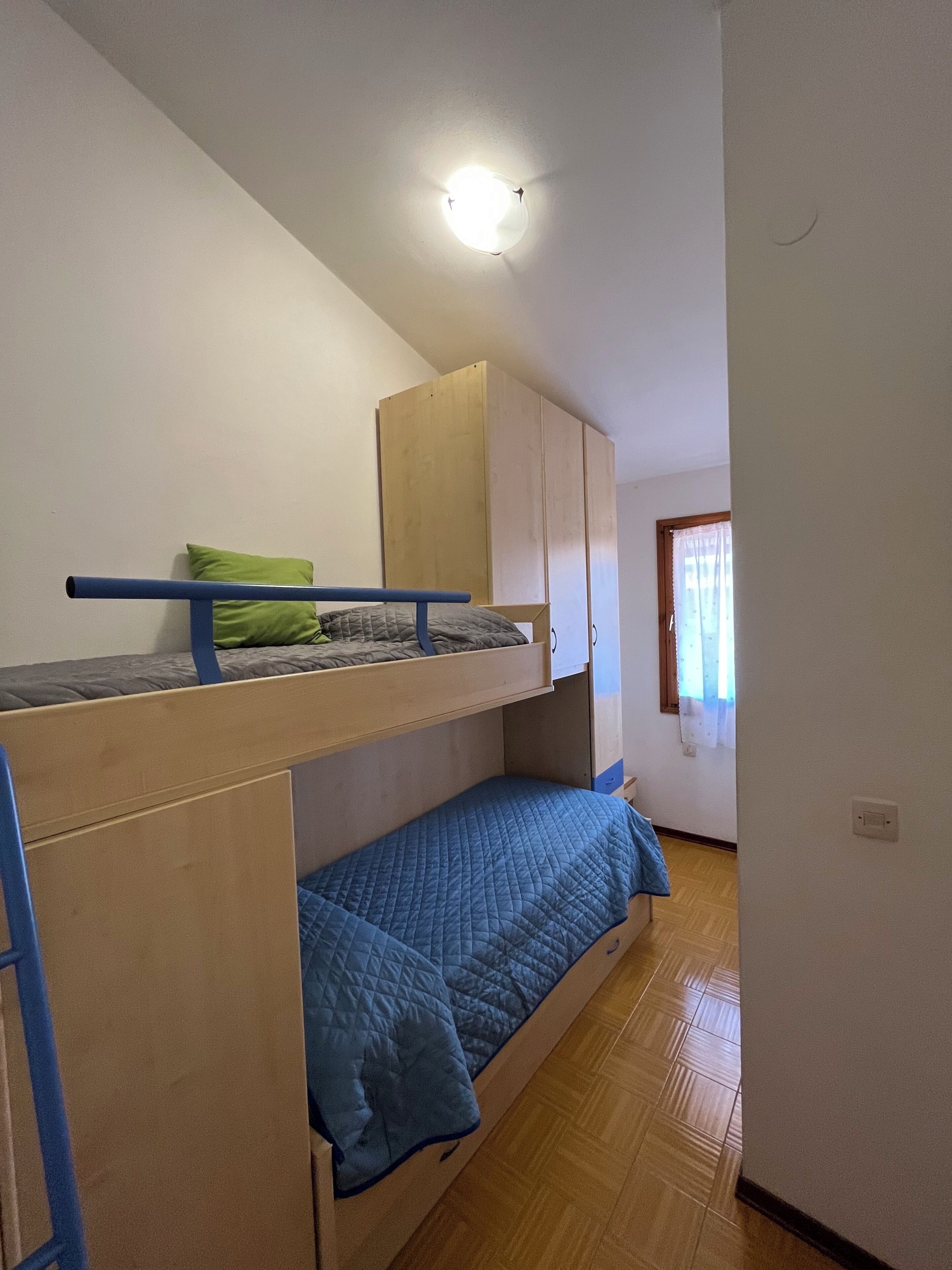 Une pièce avec des lits superposés, une armoire en bois, une fenêtre avec des rideaux et un luminaire.