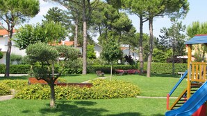 A playground with a slide and a bench in a park with trees and a building in the background. - Cozy Retreat With Garden and Pool (San Michele al Tagliamento)