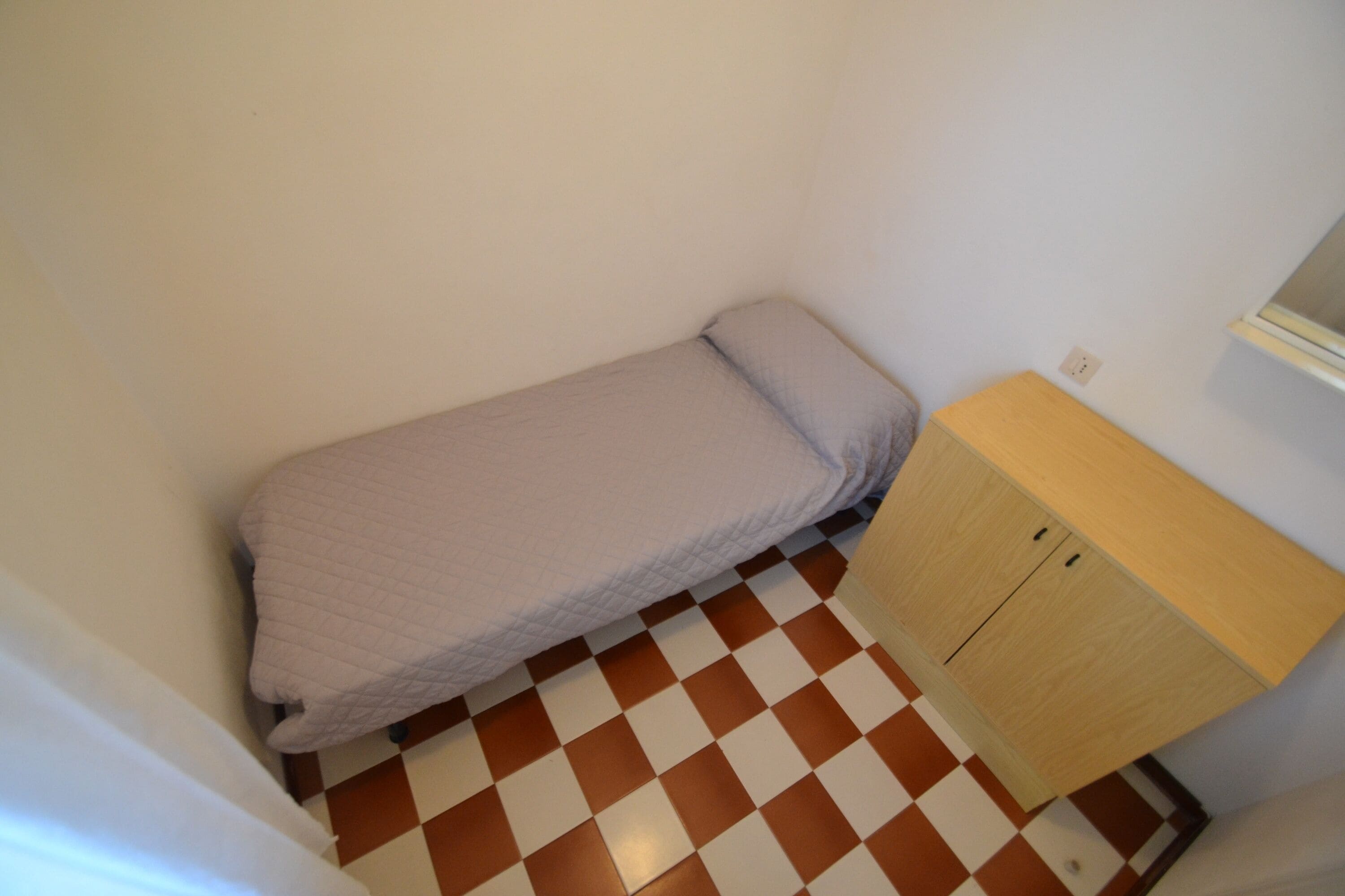 Una habitación pequeña con piso a cuadros, una cama individual cubierta con un edredón blanco y un armario de madera.
