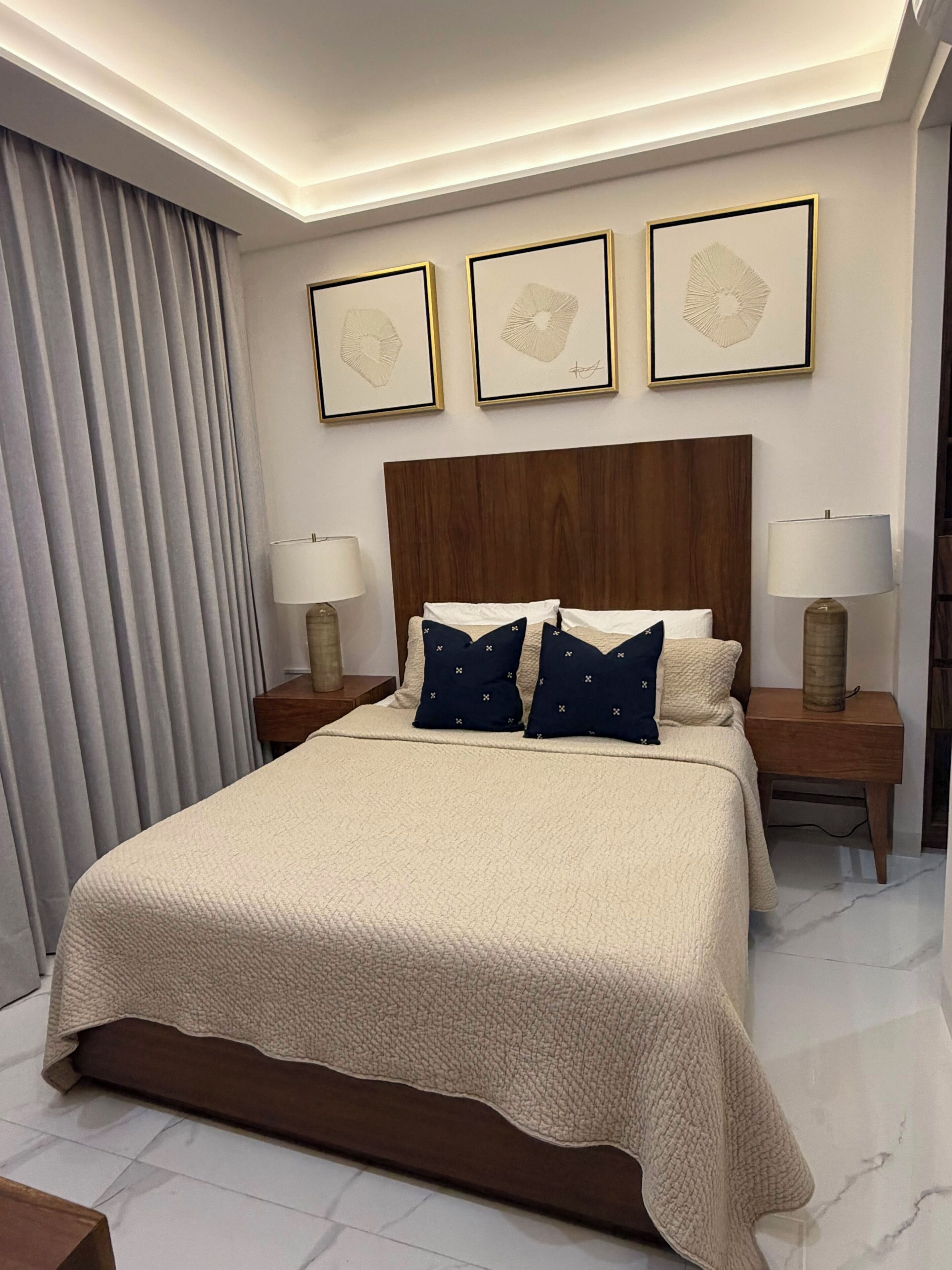 A hotel room with a large bed, wooden headboard, two bedside tables with lamps, and three framed artworks on the wall.
