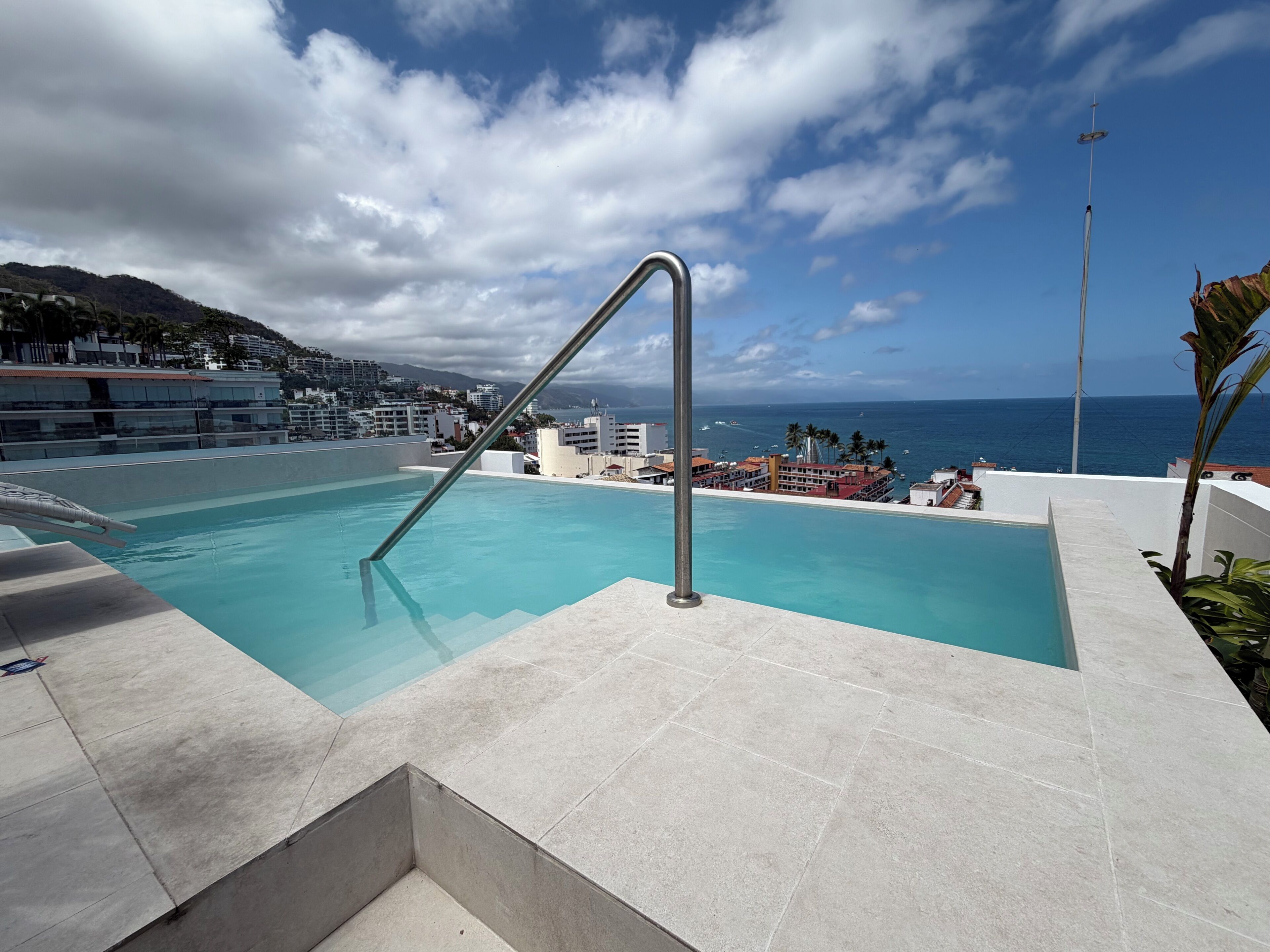 A swimming pool with a metal ladder, surrounded by a tiled deck, with a view of the ocean and buildings in the background.