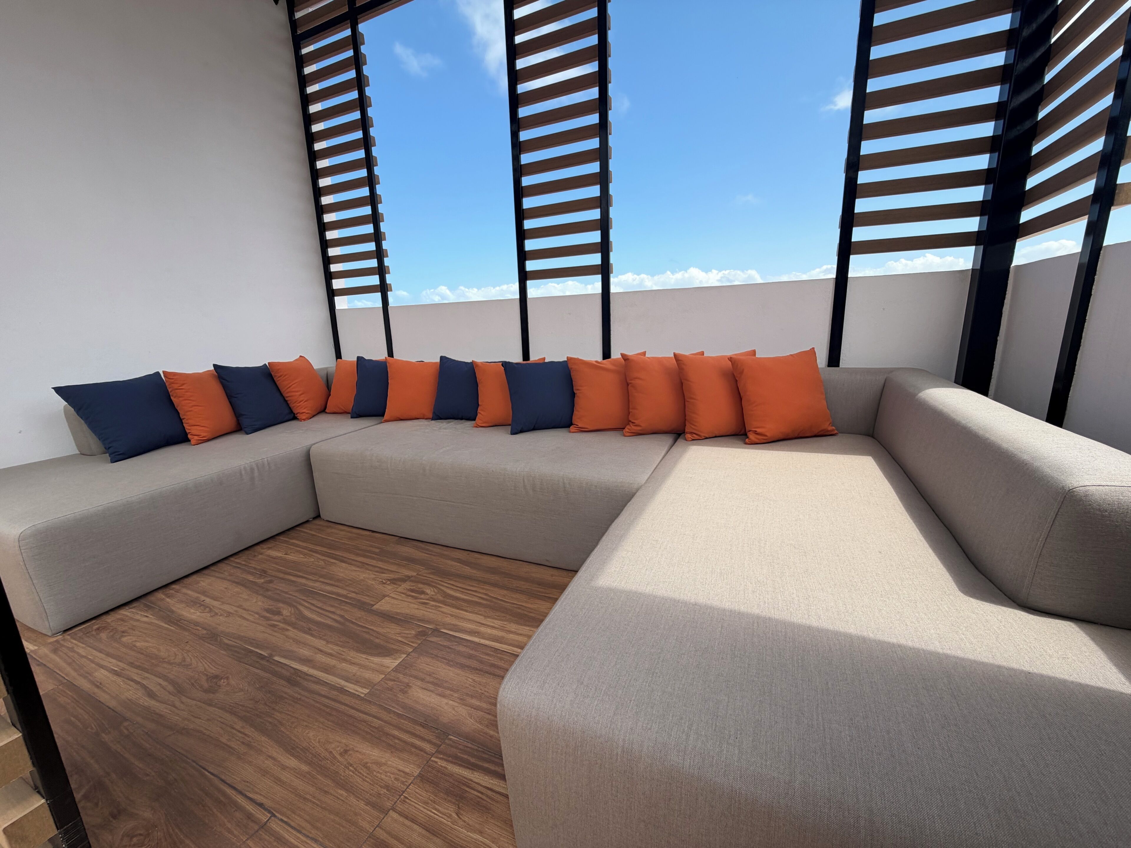 Un sofá modular con varios cojines, piso de madera y vista a través de una ventana con persianas.