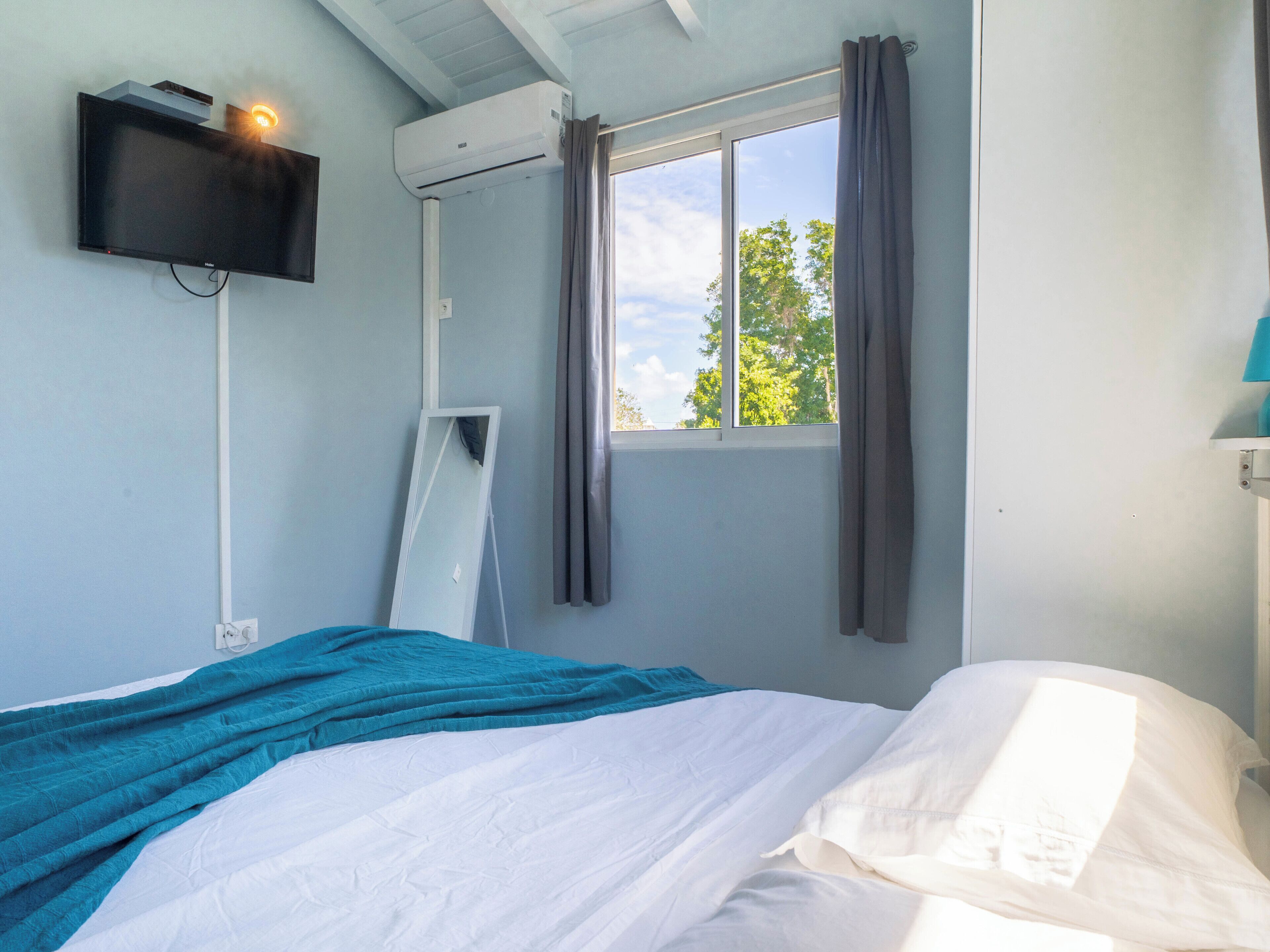 A room with a bed, a mounted TV, an air conditioning unit, a window with curtains, and a view of trees outside.