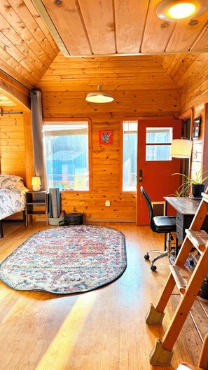 A cozy cabin interior with a wooden ceiling, a round rug, a desk with a chair, and a window with a view.